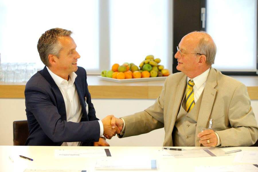 Twee volwassen heren schudden elkaar de hand na het tekenen van een overeenkomst met een fruitschaal op de achtergrond